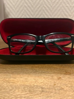 Svarta Ray-Ban Glasögon - Dessa glasögon köptes 2023 för 2900kr. De är föreskrivna och i perfekt skick.  De kommer inte med den ursprungliga Ray band Box eftersom jag inte kunde hitta den utan istället en vanlig Spec-saver box, inklusive en glasögonrengörare.  Om du har några frågor, fråga gärna, jag är aktiv oftare nu! Stilrena svarta glasögon från Ray-Ban med rektangulära bågar i plast. Diskret Ray-Ban-logga på skalmen och medföljande fodral med röd insida. Perfekt accessoar för dig som gillar klassisk och tidlös des