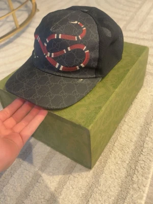 Gucci keps med  - Snygg svart keps från Gucci med klassiskt GG-mönster och en röd-vit orm på framsidan. Kepsen har mesh på sidorna och justerbar rem bak. Materialet är en mix av canvas och mesh, perfekt för en trendig streetwear-look har en vit fläck på sidan annars kepsen är mycket bra skick