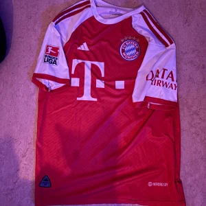 FC Bayern München matchtröja Adidas - Röd och vit matchtröja från Adidas för FC Bayern München med klubbmärke, Bundesliga-logga och Qatar Airways-tryck. Stora vita detaljer och klassiska Adidas-ränder på axlarna. Tillverkad i lätt och ventilerande material med Aeroready-teknologi.                                                                              Strl: S, men passar även M då den är lite större då de är en tränings tröja