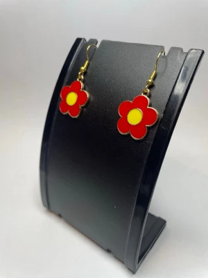 Röda blommiga örhängen i metall - Snyggas örhängen i form av röda blommor med gul mitt. Hängande modell med guldfärgade krokar och metallkant runt blomman. Perfekta för dig som vill ha en färgklick till din outfit och gillar lekfull design.