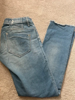 Ljusblå jeans från G-Star med rå kant - Säljer ett par ljusblå demin jeans från G-Star med snyggt slitna detaljer och rå kant nedtill. Jeansen har klassisk femficksdesign, broderad logga på bakfickan. Perfekta för en avslappnad och trendig look.