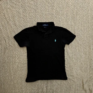 Svart pikétröja från Ralph Lauren - Klassisk svart pikétröja från Ralph Lauren med korta ärmar och turkos broderad logga på bröstet. Tröjan har krage och knappar vid halsen. Perfekt till jeans eller shorts för en clean och stilren look.