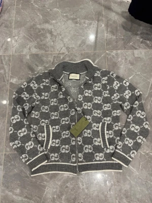 Grå ziptröja med GG-mönster från Gucci - Snygg grå ziptröja från Gucci med vitt GG-monogram över hela plagget. Tröjan har ribbade muddar, ståkrage och dragkedja framtill. Tillverkad i mjukt stickat material och har vita detaljer vid ärmslut och nederkant. Perfekt för dig som gillar exklusiv streetwear.