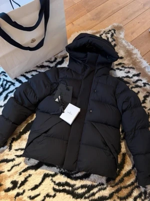 Moncler madeira  - Snygg svart dunjacka från Moncler med stor huva och breda, vadderade paneler. Jackan har diskret Moncler-logga på ärmen och präglad Moncler-text längs dragkedjan. Perfekt för kalla vinterdagar och riktigt trendig look.