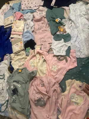 Stort paket babykläder i pastellfärger - Säljer ett stort paket babykläder i mjuka pastellfärger, med söta djur- och stjärnmotiv. Innehåller bodys, pyjamasar, mössor och omlottbodys i rosa, grönt, blått, vitt och beige. Flera plagg har tryckknappar och fina detaljer. Perfekt för dig som vill ha gulliga och bekväma babykläder.