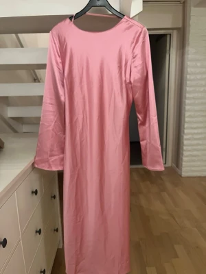 Rosa långklänning med knyt fram - Snygg rosa långklänning i glansigt material med lång ärm och v-ringning. Klänningen har ett dekorativt knyt framtill och en enkel, elegant siluett. Perfekt för dig som vill sticka ut med en feminin look. Lappen är avklippt för att den stack💕