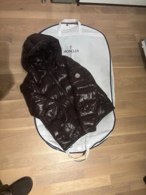 Brun dunjacka med huva från Moncler - Säljer en brun, glansig dunjacka från Moncler med huva och dragkedja framtill. Jackan har det klassiska Moncler-märket på ärmen och en unik serietecknad tvättlapp på insidan. Perfekt för kalla vinterdagar och riktigt snygg streetstil.