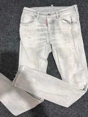 Ljusgrå distressed jeans från DSQUARED2 - Snygga ljusgrå jeans från DSQUARED2 med slitna detaljer och färgstänk. Klassisk femficksmodell med smal passform och DSQ2-logga bak i midjan. Perfekta för dig som gillar en trendig och avslappnad stil.
