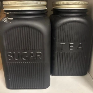 Svarta burkar för socker och te - Två stilrena burkar i svart med guldfärgade lock, märkta med 'SUGAR' och 'TEA' i upphöjd text. Burkarna är rektangulära med räfflad yta och passar perfekt för förvaring i köket. Materialet är nog glas, färgen har lossnat på vissa ställen då dom diskats i diskmaskinen. 