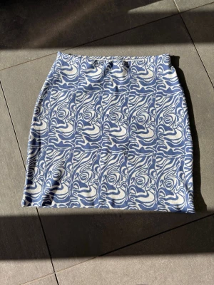 Blåvit mönstrad kortkjol - Säljer en blåvit kortkjol med unikt abstrakt mönster. Kjolen har en rak passform och är tillverkad i ett mjukt stickat material som känns bekvämt mot huden. Perfekt för dig som vill sticka ut med en cool och konstnärlig stil.