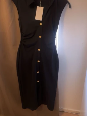 Zara Svart klänning med guldknappar - Snygg svart klänning med krage och dekorativa guldknappar längs hela framsidan. Figurnära passform och ärmlös design som ger en elegant siluett. Perfekt för dig som vill ha en stilren och chic look. Den är i storlek S (minsta storleken) aldrig använd med prislapp. 