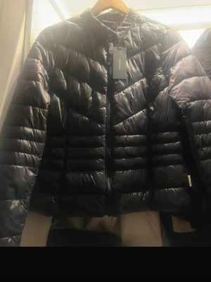 Svart dunjacka från Uniqlo - Snygg svart dunjacka från Uniqlo med quiltat mönster och hög krage. Jackan har dragkedja framtill och en lätt, puffig design som håller dig varm under kalla dagar. Perfekt för dig som vill ha en stilren och funktionell vinterjacka.