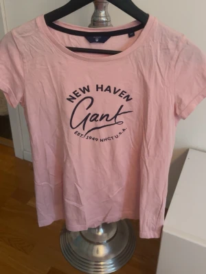 Rosa Gant t-shirt med tryck - Säljer en ljusrosa t-shirt från Gant med New Haven-tryck på bröstet. T-shirten har rund hals och korta ärmar, perfekt för en avslappnad och cool look. Materialet är mjuk bomull som känns skönt mot huden.