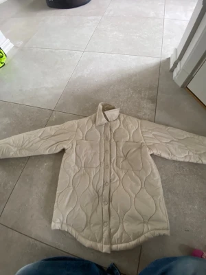 Beige quiltad pufferjacka - Säljer en beige quiltad pufferjacka med vågigt mönster och knappar framtill. Jackan har klassisk krage, långa ärmar och rundad nederkant. Perfekt för kalla dagar och riktigt skön att bära. Materialet är vadderat och ger en fluffig känsla.