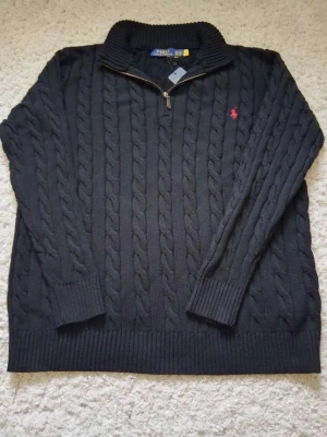 Svart kabelstickad tröja Polo Ralph Lauren - Kabelstickad svart tröja från Polo Ralph Lauren med halv dragkedja och hög krage. Tröjan har det klassiska röda Polo-logot broderat på bröstet och ribbstickade muddar vid ärmslut och nederkant. Perfekt för lager-på-lager-stil.