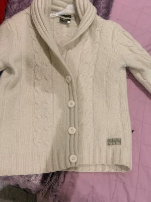 Vit kabelstickad kofta Lexington - Mysig vit kabelstickad kofta från Lexington med stora knappar framtill och sjalkrage. Koftan har långa ärmar och ribbade muddar, samt en liten logotypdetalj nertill. Perfekt för lager-på-lager och en klassisk look.