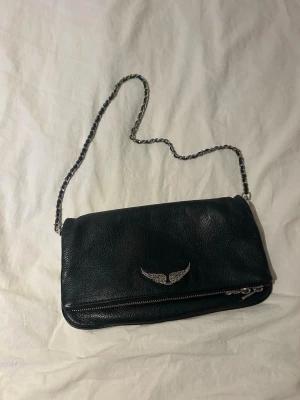 Svart kuvertväska Zadig & Voltaire - Snygg svart kuvertväska i äkta läder från Zadig & Voltaire. Väskan har en silverfärgad kedjerem och ett coolt vingemblem framtill. Dragkedja längst ner på framsidan och innerfack för småsaker. Perfekt accessoar för att lyfta din outfit. Lite sönder på kedjans läder se bilden