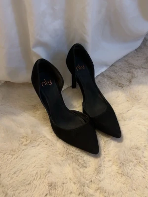 Svarta pumps  - Säljer ett par klassiska svarta pumps från NLY Shoes med spetsig tå och hög klack. Skorna har en stilren design med öppen insida och är tillverkade i ett mockaliknande material. Perfekta för att lyfta vilken outfit som helst.
