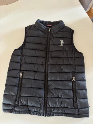 Svart dunväst från U.S. Polo Assn. - Snygg svart dunväst från U.S. Polo Assn. med quiltad design och dragkedja framtill. Västen har två praktiska fickor med dragkedja och en broderad logga på bröstet. Perfekt för lager på lager och ger en sportig look.