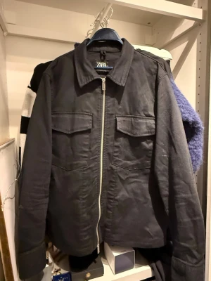 Svart overshirt från Zara - Stilren svart overshirt från Zara med dragkedja framtill och två bröstfickor med lock. Klassisk krage och raka ärmar ger en clean look. Perfekt att bära över en t-shirt för en avslappnad men snygg stil.