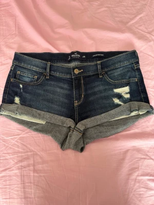 Mörkblå denimshorts från Hollister - Snygga mörkblå denimshorts från Hollister med slitna detaljer och uppvikta benslut. Klassisk femficksmodell med knapp och dragkedja framtill. Perfekta för varma dagar och ger en avslappnad vibe.