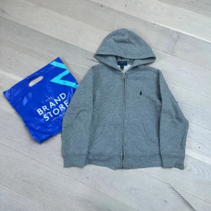 Grå hoodie från Polo Ralph Lauren - Klassisk grå hoodie med dragkedja från Polo Ralph Lauren. Tröjan har huva, två fickor framtill och den ikoniska lilla loggan broderad på bröstet. Perfekt för en chill och avslappnad stil. Tillverkad i mjukt bomullsmaterial.