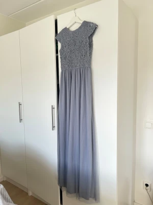 Ljusblå långklänning med spetsdetaljer - Elegant ljusblå långklänning från Bubbleroom med vackra spetsdetaljer och broderad överdel. Klänningen har v-ringning i ryggen och ett flytande, tunt kjolparti. Perfekt för speciella tillfällen och studentfirande. Prislapp sitter kvar. 