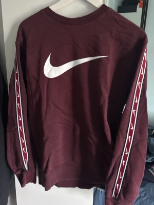 Vinröd Nike sweatshirt med logga - Snygg vinröd sweatshirt från Nike med stor vit Swoosh-logga på bröstet och vita loggor längs båda ärmarna. Tröjan har rund halsringning och ribbade muddar. Perfekt för en sportig och avslappnad stil.