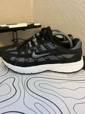 Nike P-6000 svart/grå sneaker - Nike P-6000 sneakers i svart och grå mesh med svarta syntetiska överdrag och vit chunky sula. Ikonisk Swoosh-logga i silver på sidan och sportig design med snörning. Perfekt för dig som gillar retrovibbar och streetwear.