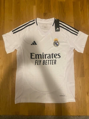 Real Madrid matchtröja Adidas vit S - Säljer en vit Real Madrid matchtröja från Adidas med svarta detaljer och klubbmärke på bröstet. Tröjan har V-ringning, tre svarta ränder på axlarna och Aeroready-material som andas. Emirates Fly Better-logga på framsidan. Perfekt för fotbollsträning eller match.