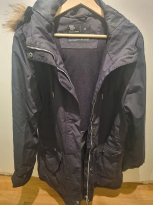 Svart parkas från Neomondo XL - Svart parkas från Neomondo i storlek XL. Jackan har dragkedja, stora fickor framtill och en huva med fuskpälsdetalj. Perfekt för kalla dagar med sitt tjocka material och höga krage. Enkel och stilren design som passar till det mesta.