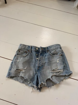 Ljusblå slitna jeansshorts - Säljer ett par ljusblå jeansshorts med hög midja och råa, fransiga kanter. Shortsen har slitningar framtill, 