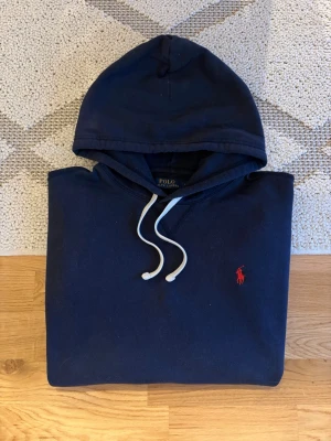 Mörk Marinblå Polo Ralph Lauren Hoodie - Hej! 👋 Säljer en riktigt snygg Polo Ralph Lauren hoodie i storlek L. 💎 Mörk marinblå färg (en klassisk och lättmatchad nyans!) ✨ Mycket fint skick! 🎉 Sitter riktigt bra (Storlek L) 👕 Den klassiska röda broderade pony-loggan på bröstet! 🐎 Alla tvättlappar sitter kvar (se bilder). ✅ Fraktar snabbt! 🚚📦 Hör av er vid frågor! 💬💯