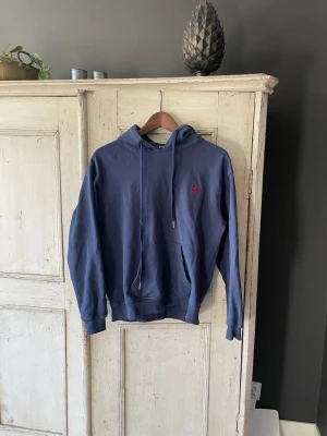 Mörkblå hoodie från Polo Ralph Lauren - Snygg mörkblå hoodie från Polo Ralph Lauren med klassisk huva och dragsko. Har en känguruficka framtill och den ikoniska röda loggan broderad på bröstet. Tillverkad i mjuk bomull som känns skön mot huden. Perfekt för en avslappnad och stilren look. Ai gjorde beskrivning