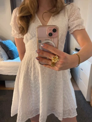 Vit kort klänning  - Superfin vit kort klänning med v-ringning och korta puffärmar. Klänningen har ett diskret prickigt mönster i tyget och knappar framtill. Perfekt för sommarens alla tillfällen och ger en romantisk vibe. Passar S/M och är använd en gång!