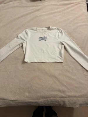 Vit croppad långärmad topp Nike - Snygg vit croppad långärmad topp från Nike med tryckt logga framtill. Perfekt att styla med jeans eller kjol för en sportig vibe. Mjuk och skön bomullskänsla, passar dig som gillar streetwear.