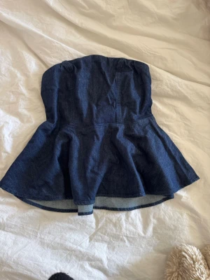 Mörkblå denim tubtopp med vidd - Säljer en snygg mörkblå tubtopp i denim med utsvängd nederdel. Toppen är axelbandslös och har en smockad baksida för extra komfort. Perfekt att styla med jeans eller kjol för en cool look.