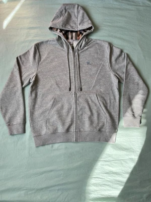 Grå zip-hoodie från Burberry - Snygg grå hoodie från Burberry med dragkedja och klassiskt rutigt foder i huvan. Broderad logga på bröstet och två stora fickor framtill. Perfekt för en stilren och avslappnad look. Mjuk bomullsblandning och justerbar huva med snören.