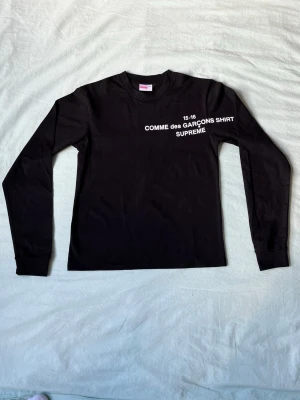 Svart Supreme x Comme des Garçons långärmad - Svart långärmad t-shirt från Supreme och Comme des Garçons med vit text på bröstet. Klassisk rund hals och rak passform. Perfekt för dig som gillar streetwear och vill ha något stilrent men ändå unikt.