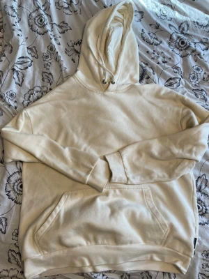 Krämvit hoodie med magficka - En stilren krämvit hoodie med huva och dragsko. Hoodien har en stor magficka framtill och ribbade muddar vid ärmslut och nederkant. Tillverkad i mjukt bomullsliknande material som känns skönt mot huden. Perfekt för en avslappnad look.