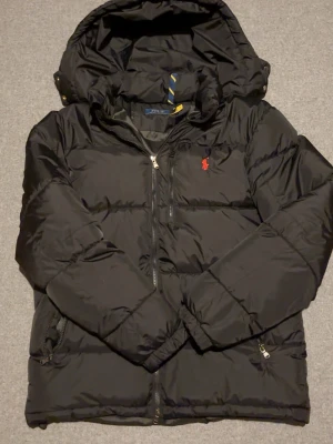 Svart pufferjacka från RL - Snygg svart pufferjacka från RL med huva och dragkedja framtill. Jackan har det klassiska röda Polo-logot på bröstet och praktiska fickor med dragkedja. Perfekt för kalla dagar och riktigt stilren look.