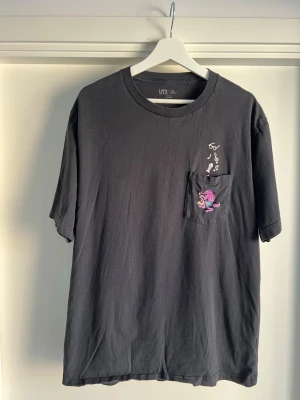 Svart t-shirt med ficka och brodyr - Svart limited edition t-shirt från UT med kort ärm, bröstficka och broderad design med musiknoter och en färgglad figur på fickan. Avslappnad passform och rund halsringning. Perfekt för en chill och stilren look.