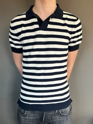 Randig pikétröja i marinblått och vitt - Klassisk pikétröja med marinblå och vita horisontella ränder. Tröjan har krage, korta ärmar och ribbade muddar. Perfekt för en avslappnad och stilren look. Materialet känns mjukt och bekvämt mot huden.