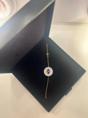 Guldarmband med öga  - Snyggt armband i guldton med en kedja, dekorerat med ett öga i mitten omgivet av små vita pärlor och ett litet kors på sidan. Perfekt accessoar för dig som gillar unika detaljer och symbolik.