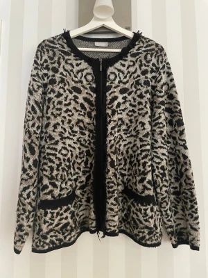 Leopardmönstrad kofta med dragkedja - Säljer en snygg leopardmönstrad kofta i svart och beige med dragkedja framtill. Koftan har två fickor och fransdetaljer längs kanterna. Perfekt för dig som gillar djurmönster och vill ha en unik look.