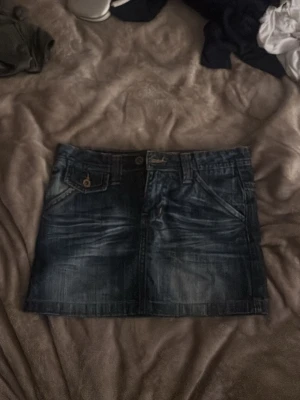 Mörk jeanskjol med fickor - Snygg mörkblå jeanskjol i kort modell med klassiska fickor fram och bak samt bälteshällor. Kjolen har en knapp- och dragkedjestängning framtill och coola slitningar för en trendig look. Perfekt att matcha med en t-shirt eller hoodie.