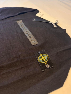 Svart stickad tröja Stone Island - Snygg svart stickad tröja från Stone Island med logotyp i relief på bröstet och den klassiska patchen med kompass på ärmen. Tröjan har ribbade muddar och rund hals. Perfekt för dig som vill ha en stilren och exklusiv look.