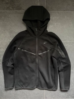 Svart Nike Tech Fleece hoodie - Säljer en svart Nike Tech Fleece hoodie med dragkedja framtill och huva. Hoodien har en diskret Nike-logga på vänster bröst och en ficka med dragkedja på ärmen. Tillverkad i mjukt och stretchigt material som ger en clean och sportig look. 