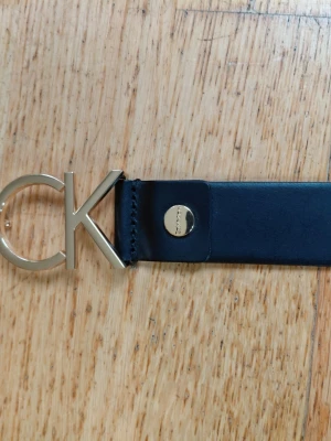 Svart skärp från Calvin Klein - Stilrent svart skärp från Calvin Klein med bred rem i skinn och ett stort guldfärgat CK-spänne framtill. Perfekt för att lyfta vilken outfit som helst med en snygg logodetalj. Jag har skapat 2st hål enligt bild.