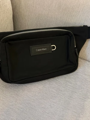 Svart shoppingväska från Calvin Klein - Snygg svart shoppingväska från Calvin Klein i slitstarkt tygmaterial. Väskan har två stora fack med silverfärgade dragkedjor och ett tydligt logomärke framtill. Perfekt för dig som vill ha en stilren och praktisk väska med plats för det viktigaste. Går att använda som Mag/Axelväska